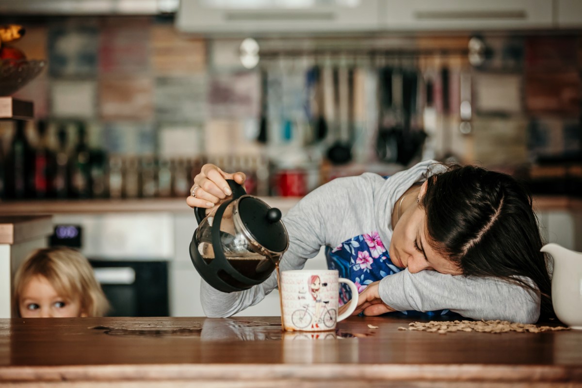 O femeie obosită stă întinsă pe tejgheaua din bucătărie, vărsând cafeaua lângă o cană, cu un copil lângă ea.