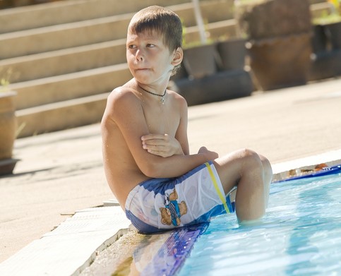 Băiatul stă lângă piscină, cu picioarele în apă. Îi este frig.